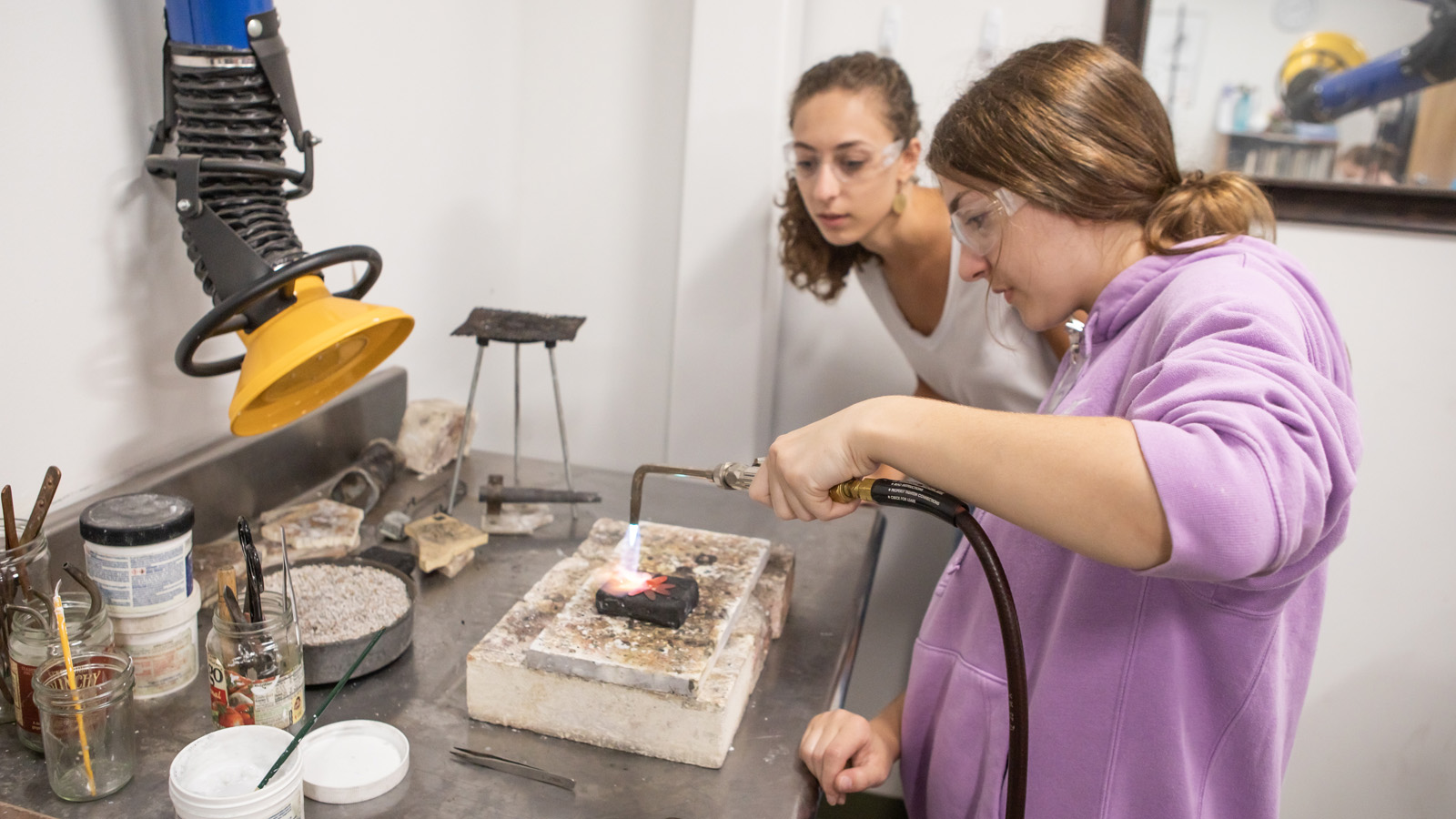 a youth with a torch firing her stamped jewelry, and another youth looks on.