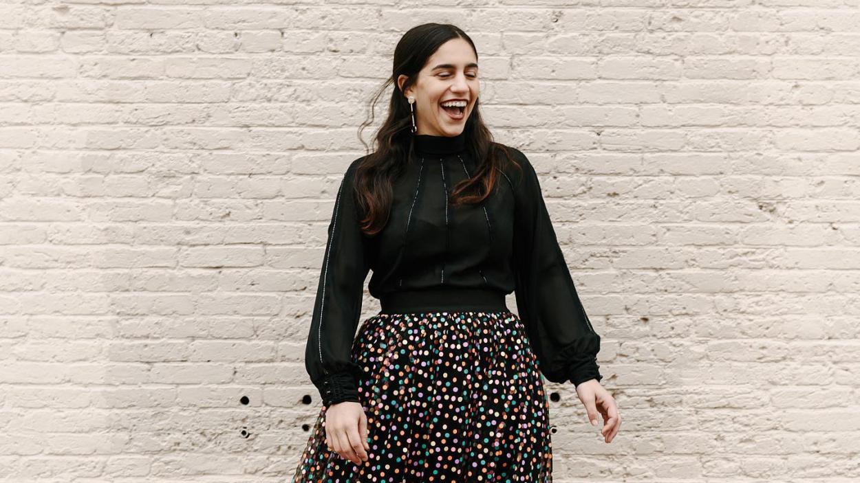 A light skinned woman wearing a black high necked blouse with full sleeves, and a black shirt with small multi-colored polka dots laughs and stands in front of a brick wall painted white