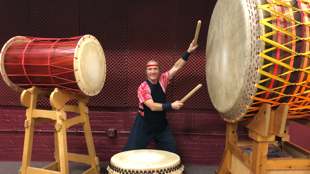 A light skinned man wearing a black bib vest over a red tie dyed shirt, with a red headband holds two large drum sticks and stands ready to strike the oversized drums on either side and in front of him.