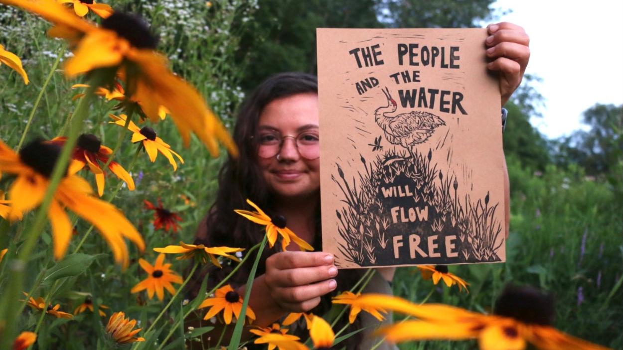 a photograph of a woman with long brown hair and glasses sitting in a field of black eyed susans holding a print that says The People and the water will flow free with illustrations