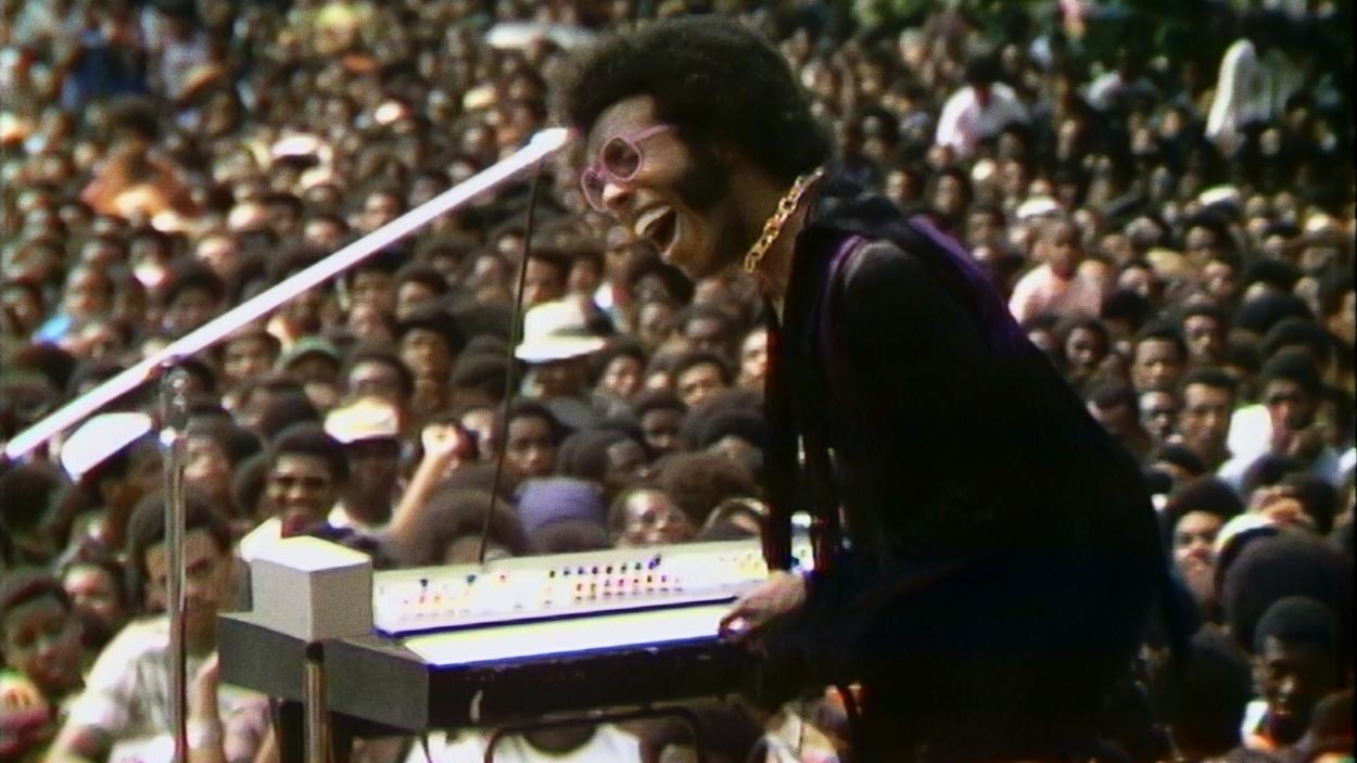 A photograph of sly stone, a dark skinned man wearing sunglasses singing into a microphone, with a crowd of people in the background
