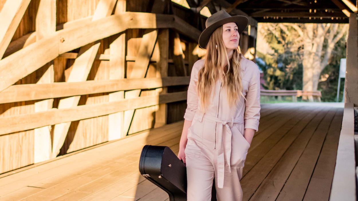 A light skinned woman with wavy blonde hair walks along a covered bridge carrying a guitar case, wearing a light pink jumpsuit and black wide brimmed hat