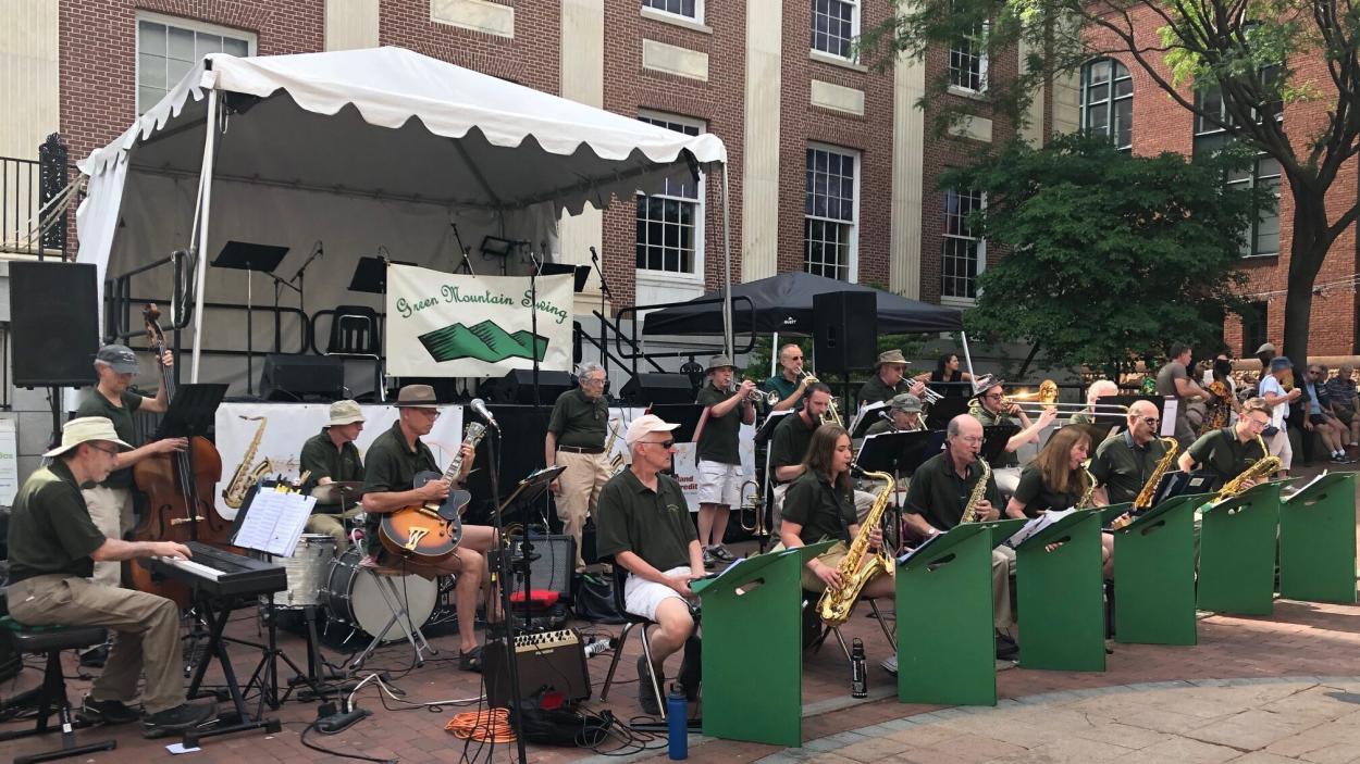 An 18 piece jazz band plays outdoors on the brick surface of the Church Street Marketplace, with a white tent over a raised stage and Burlington's City Hall, a brick and white stone building behind them.