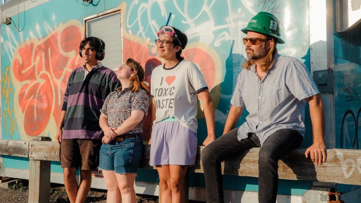 Three light skinned men and one light skinned woman stand against a wall covered with colorful graffiti, bathed in golden hour sunlight. One man wears a green hard hat, one a snorkel mask and a tee shirt that says "I love toxic waste" and the woman has a small plush horse sitting on her shoulder.