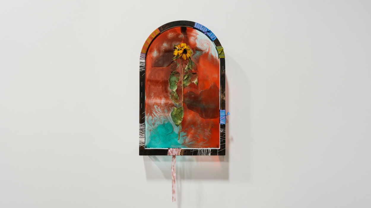 A photograph of a wall hanging sculpture by artist Lydia Kern, called Double Sorrow, Double Joy. The sculpture is shaped like an arched window and is made of colorful acrylic and flowers