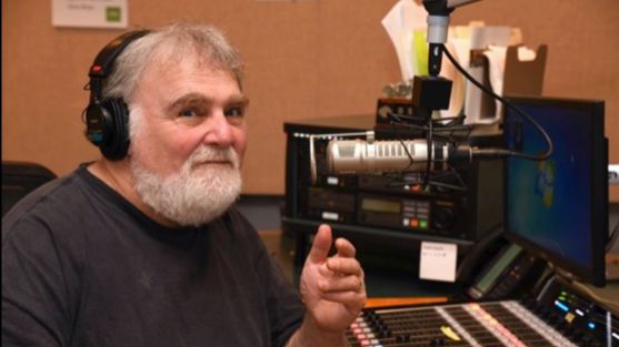 A photograph of Herb Lockwood, a light skinned man with a silver beard and hair sitting at a mixing board, in front of a microphone