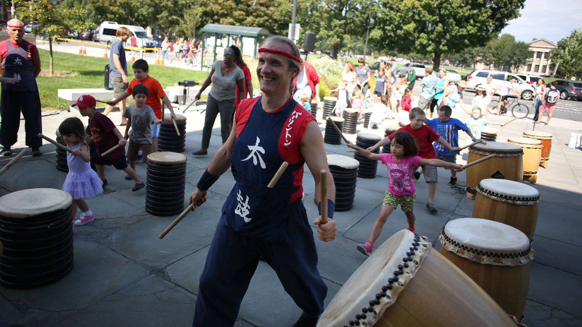 A man wearing a navy blue and red shirt with white Japanese characters leads a group of children striking a large drum 