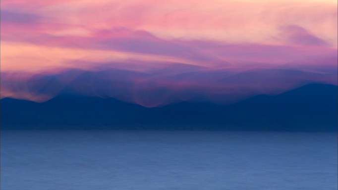 a painterly abstract photography of blue lake, with darker blue mountains and lavender and peach sky filled with wispy clouds