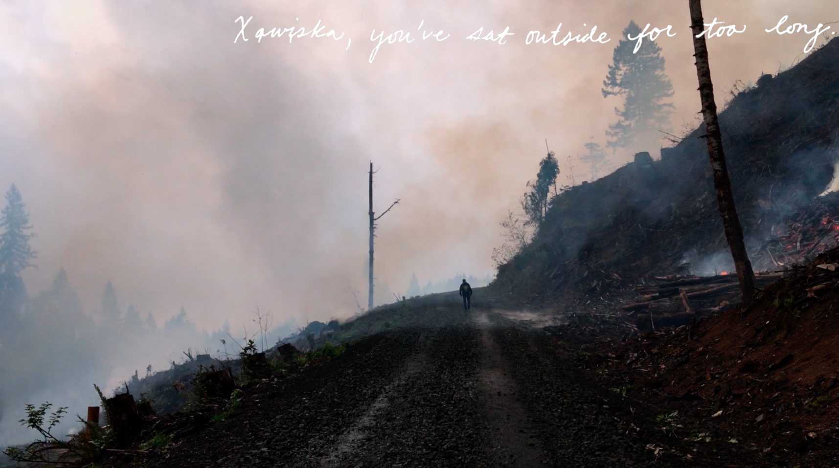 a still from a film that shows a man walking down a dirt road amidst a smoking landscape that has recently been burned with white handwriting that says &quot; Xwįska, you've sat outside too long&quot;