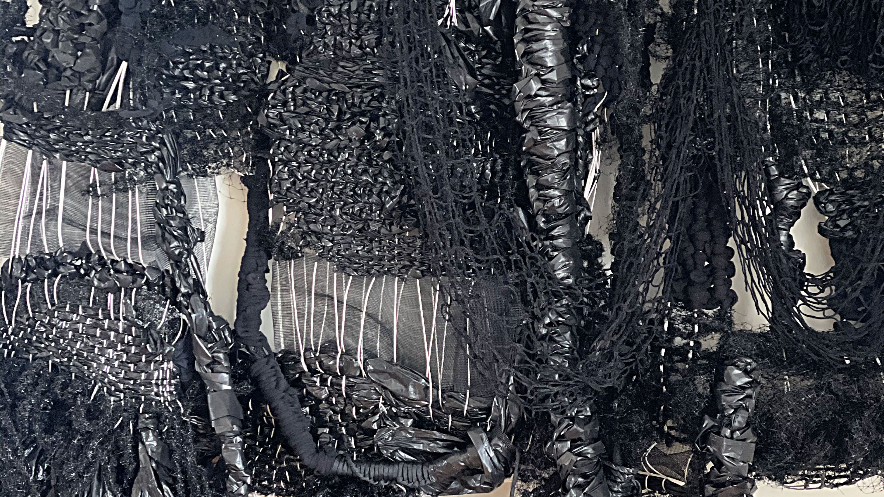 A close up of a textile sculpture that hangs on a white wall, consisting of knotted black cotton, plastic, and thread interwoven in varying patterns