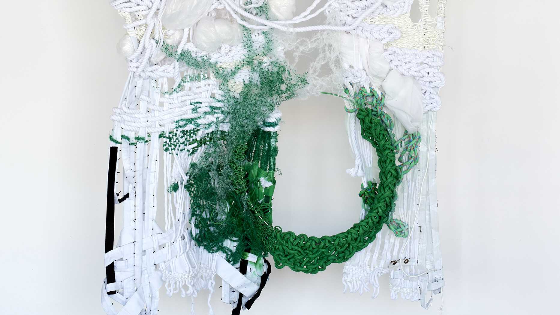 A textile sculpture that hangs on a white wall, consisting of knotted white cotton, plastic, and thread interwoven with rich green pieces that form a circle of negative space