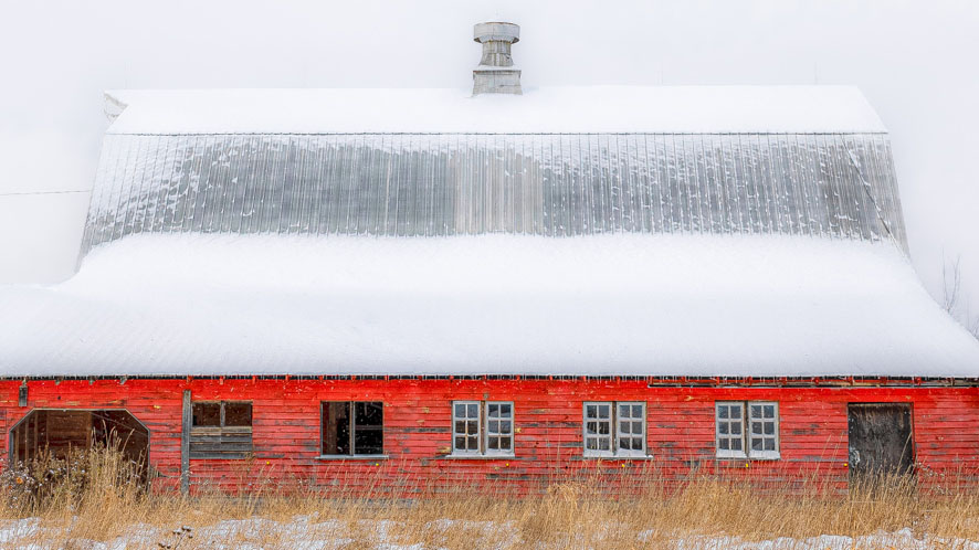 a red barn with a silver hipped standing seam metal roof that is lightly dusted with snow against a background of hazy gray