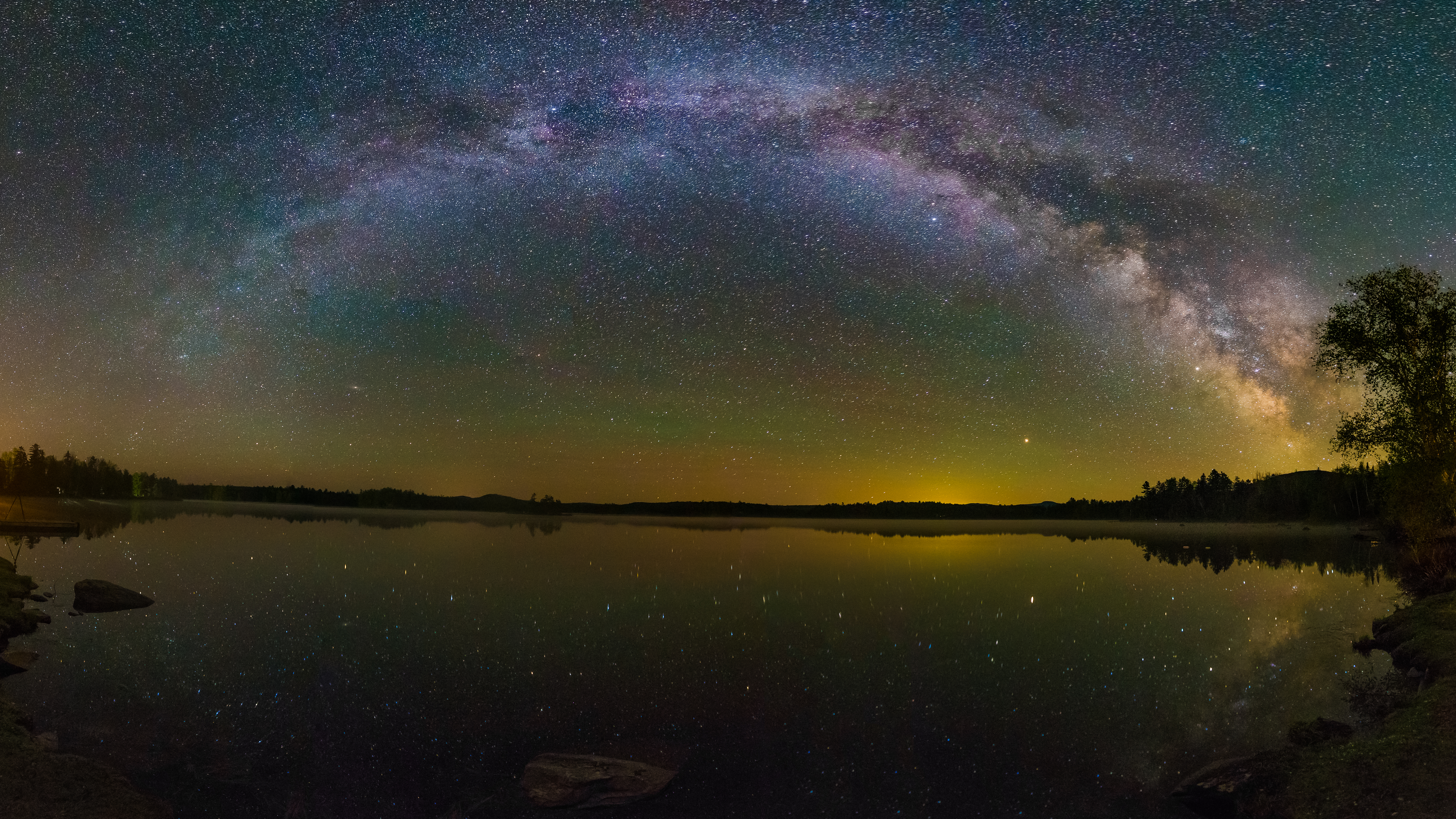 A night time photograph of a lake as the last daylight fades over the horizon under a greenish black sky dense with stars and purple northern lights