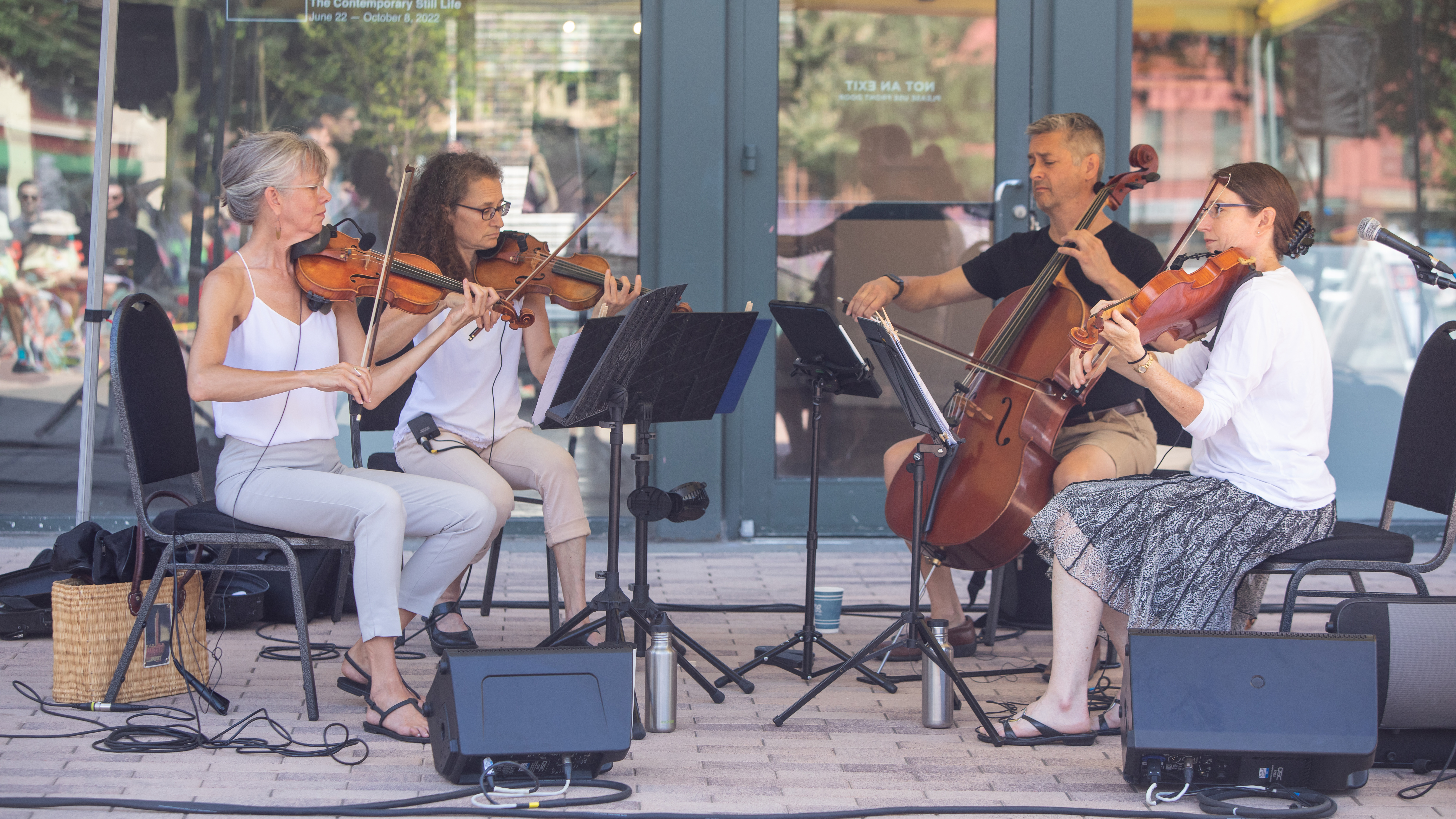 A string quartet of three light skinned women and one light skinned man play on a shady patio. The women each play a violin and the man plays a cello. Large glass windows and a door behind them captures their reflection.