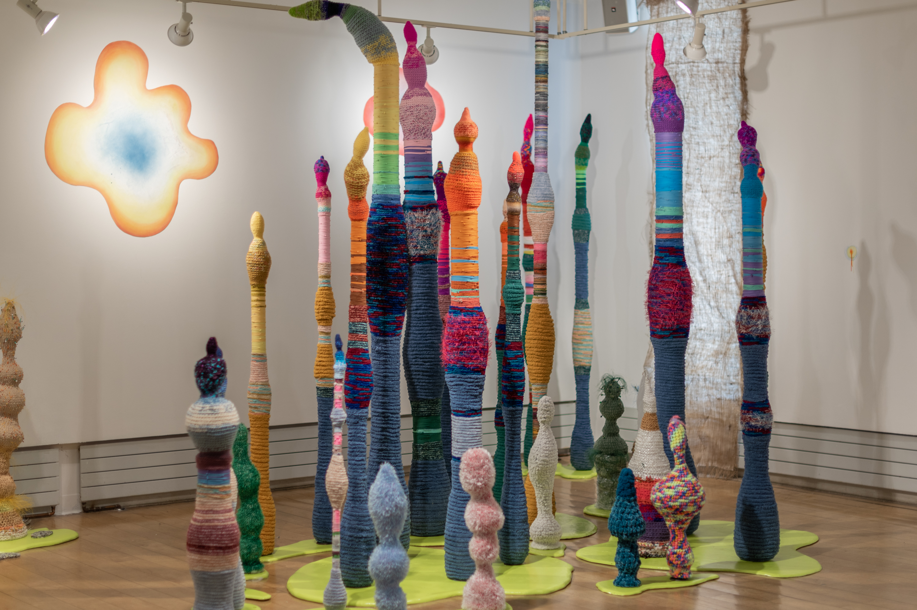 A gallery filled with tall, bulbous, linear multi colored sculptures with textured surfaces made from crocheted yarn. The sculptures rise from lime green bases in abstract blobby shapes. An abstract curvy blob that is blue in the center, fading to orange and yellow at the edges hangs on the wall in the background