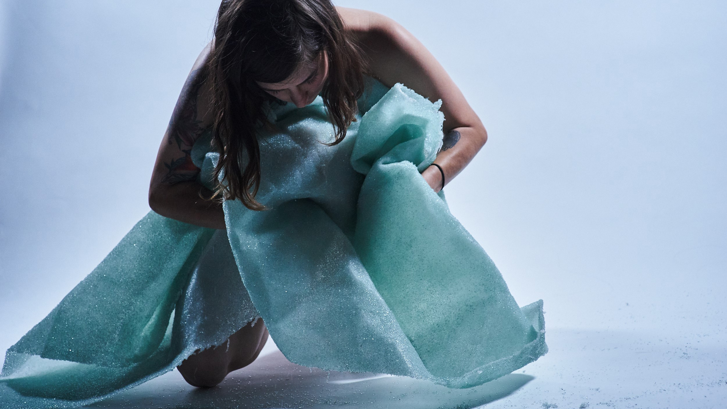 A naked light skinned woman embraces a green, crumpled glass blanket against a white background