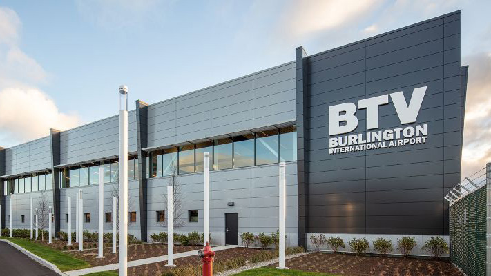 a digital illustration of a a silver and grey building that says BTV Burlington International Airport in white lettering, with white poles and small bushes in front