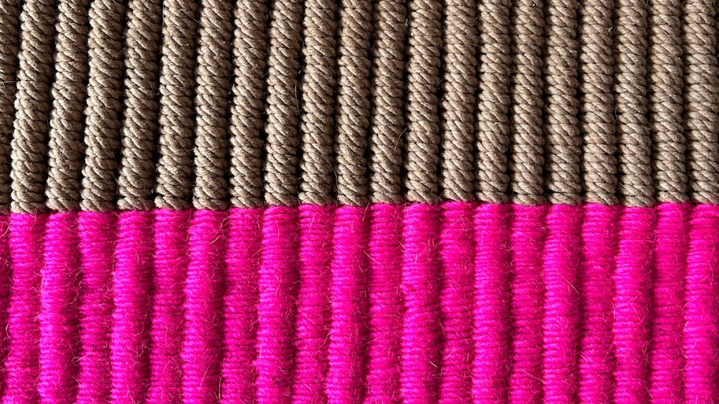 a photograph of a close up of a piece of textile art comprised of tightly woven ribs of thread. The top half are light brown and appear to be made of jute and the bottom half are bright magenta.