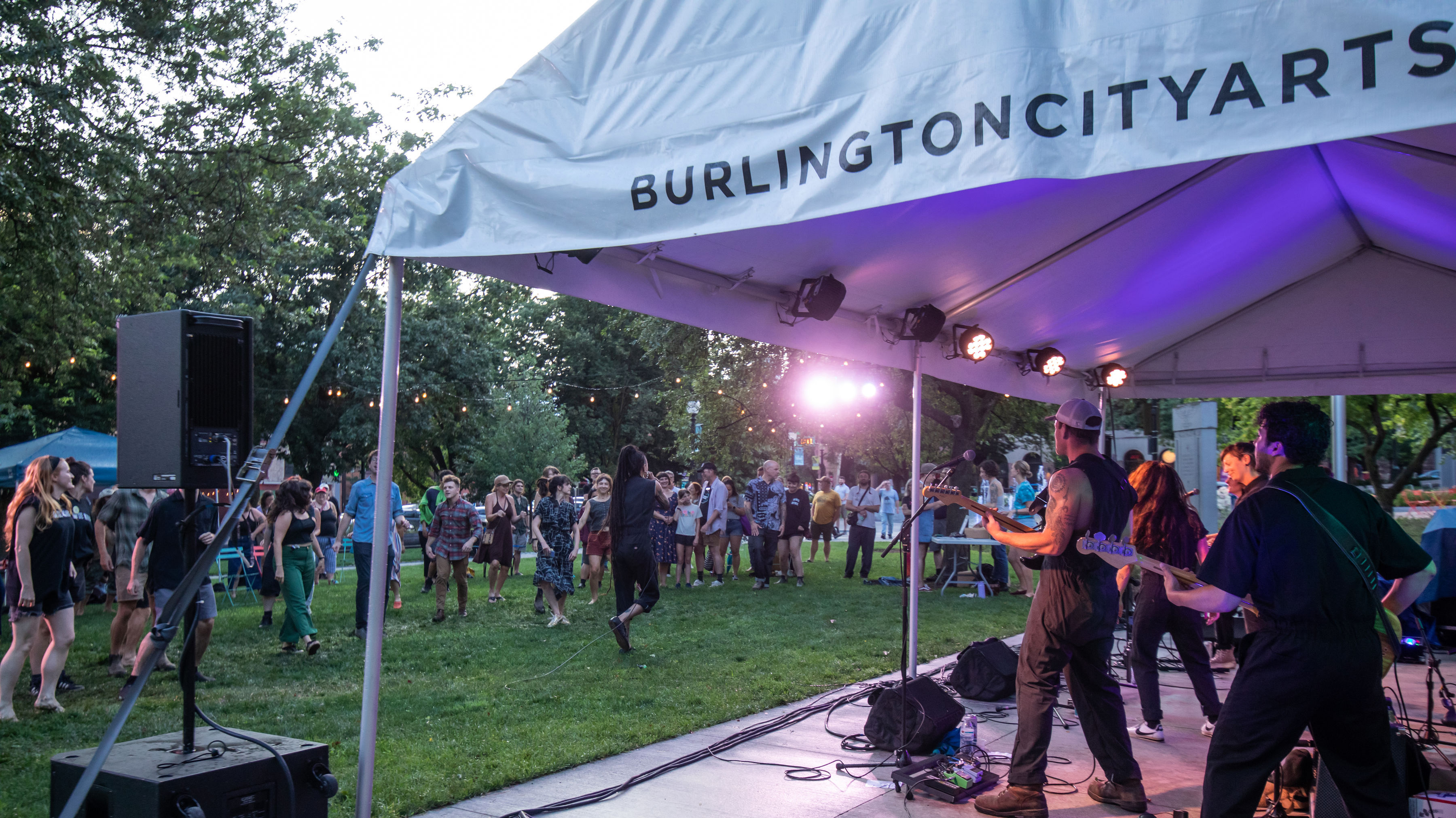 A photograph of an outdoor concert. A band with serveral light skinned men and women wear black jumpsuits and play guitar and bass under a white tent that says burlingtoncityarts.org. A dark skinned man with dreadlocks that go all the way down his back, also wears a black jump suit and sings into a microphone out in the crowd, who dance on green grass. 