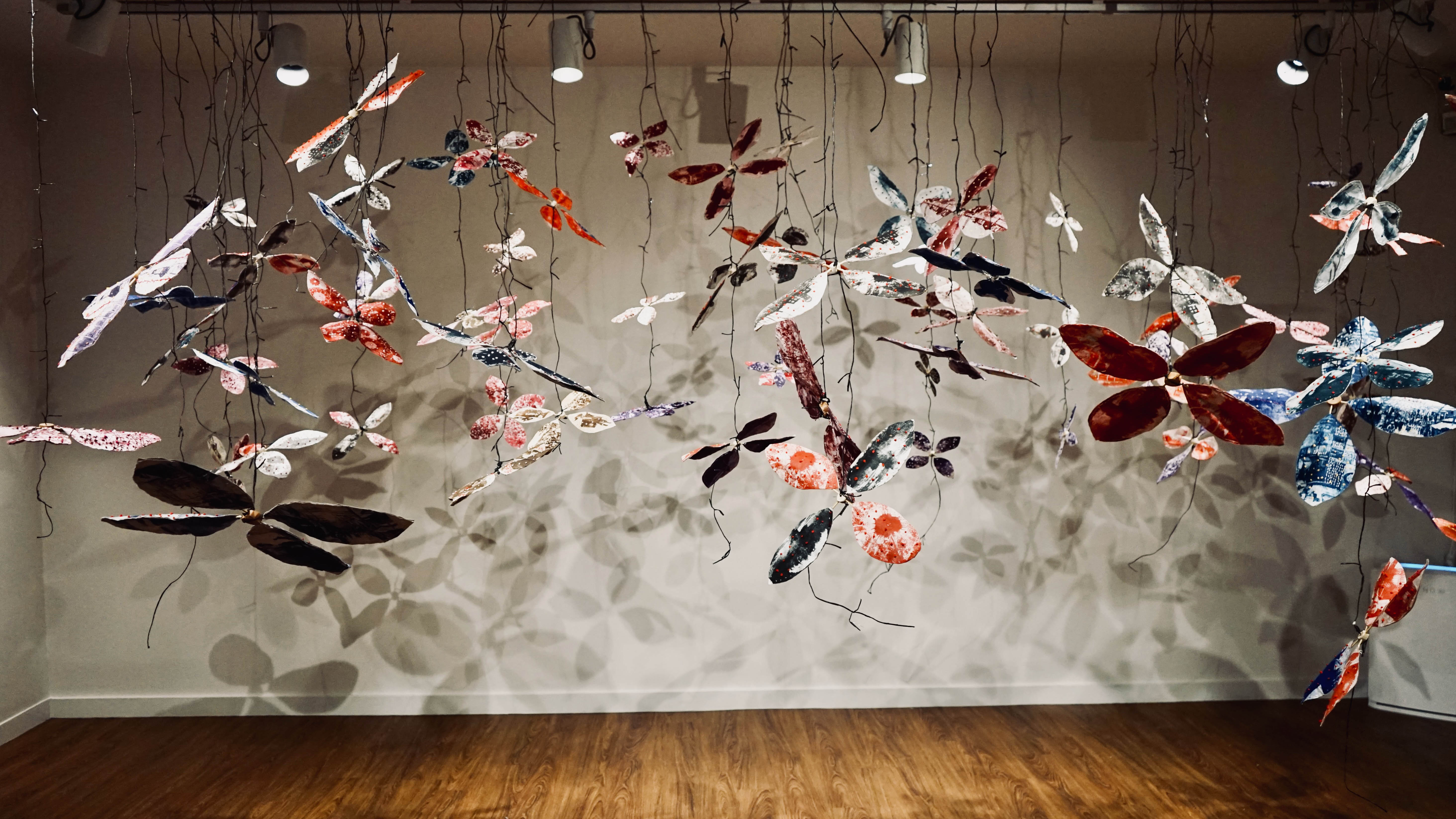 A photograph of a sculptural installation featuring many paper forms that resemble flowers or dragonflies suspended from the ceiling. They throw shadows on the white wall behind them.