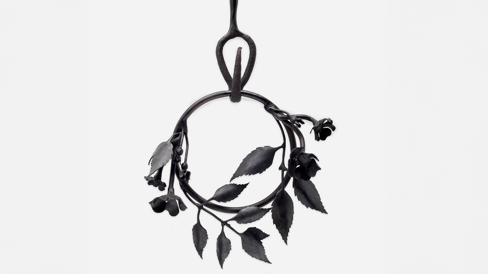 A photograph of a detail of a black steel sculpture that consists of a ring with leaves, flowers and vines twined around hanging from a hook a ring