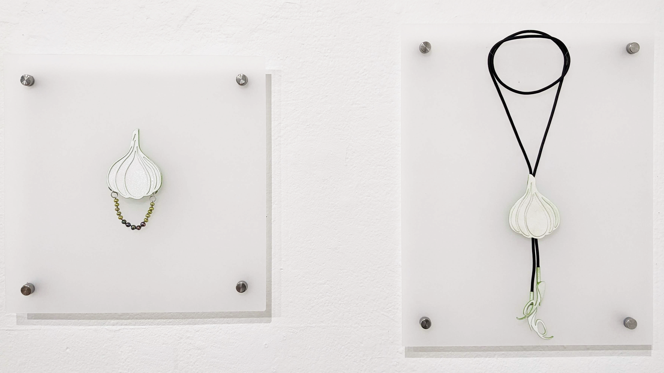 A photograph of two pieces of jewelry, mounted on a white wall. On the right is a brooch with a white bulb of garlic with a delicate chain looped to the bottom side. On thee right is a bulb of garlic hanging from a brown leather string looped around several times