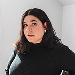 A headshot of a Leslie Sainz, a Cuban woman with medium toned skin, dark hair that falls just below her shoulders, and dark eyes. She wears a black turtleneck