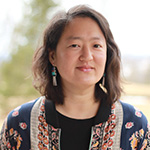 A headshot of Sarah Audsley, a Korean American woman with medium toned skin and dark, wavy, shoulder length hair. She wears a navy patterned top and long gold and green earrings
