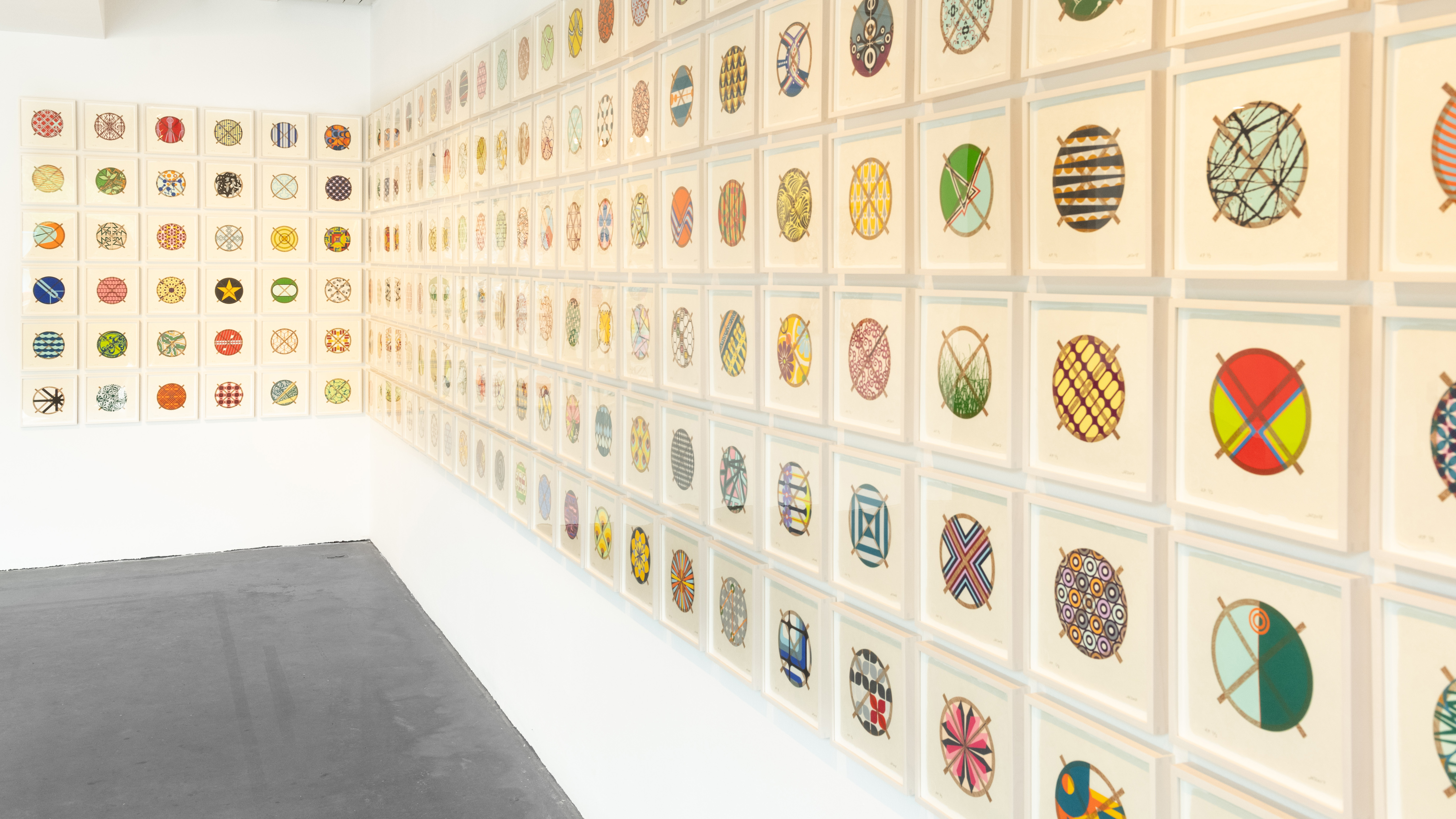 a photograph of an installation in the BCA Center gallery by Jacob Hashimoto made up of hundreds of framed circular, colorful prints, hung in a grid that wraps around a corner of the gallery wall