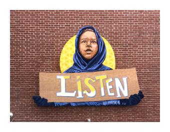 A mural depicting a young black boy wearing a navy blue hoodie, holding a sign that says "Listen" with the sun shining behind him.