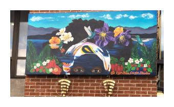 A mural that shows a black woman's face from the eyes up, with tropical flowers in her hair, and mountains and a lake in the background. Large gold earrings hang below the mural.