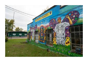 a mural painted on a brick building depicting three black teenagers with elaborate purple wings, standing in a colorful field of sunflowers