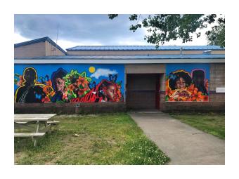 A mural mounted on a low, cement building that depicts dark skinned adolescent boys and girls, outlined in vibrant colors against a bright blue sky background with large, stylized orange, pink and red flowers