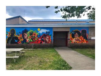 A mural mounted on a low, cement building that depicts dark skinned adolescent boys and girls, outlined in vibrant colors against a bright blue sky background with large, stylized orange, pink and red flowers 