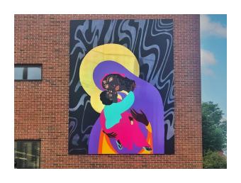 A mural mounted on a brick building depicts a dark skinned madonna and child, with the mother wearing a purple, pink and yellow robe and the child held in her arms, reaching for her bowed head wearing an aqua tunic. The background is black and grey marbleized swirls