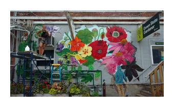 a mural of lush hyper realistic flowers in reds, yellows, pinks, and greens on the side of a grey brick building