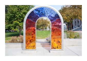 Close up of a colorful acrylic art installation at the edge of a city park with trees in the background.
