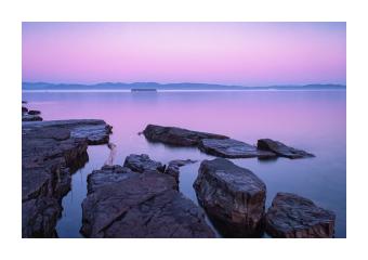 Photograph of a rocky water's edge at sunrise in purples and blues. 