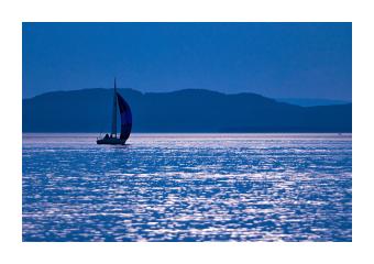 Photograph of a lake surface with mountains in the background and a sailboat to the far left of the water. The photograph is in shades of blue. 
