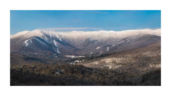 Panoramic photograph of a mountainscape dotted by browns and whites with a blue sky. 