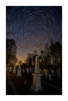 Photograph at a cemetery pointed at the sky where star trails spiral into one another in blues and yellows. 