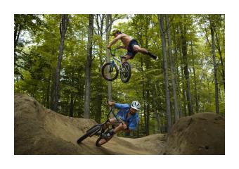 Photography of two figures biking on a wooded path where one is above the other in the air. 