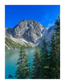 Photograph of a view of a mountain and lake with pinetrees in the foreground.