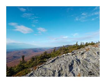 Photograph from the top of a mountain looking across at the sky and down at the forest, blues and browns.