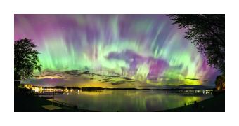 Photograph depicting the aurora borealis on a lakefront, where the sky is pale purple, blue, and seafoam green.  