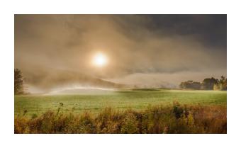 Photograph of a foggy sunrise over a field and hills. 
