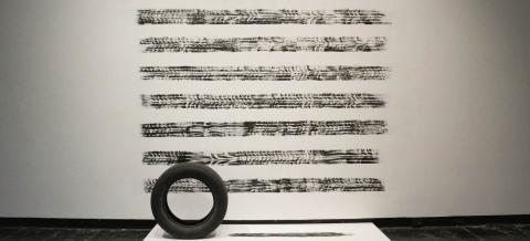 A photograph of a sculptural installation featuring a black tire on a low white platform, with 8 horizontal stripes of black tires tracks on the wall behind.