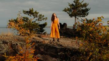 Artist standing in an orange linen pantsuite on rocky cliff in front of lake champlain