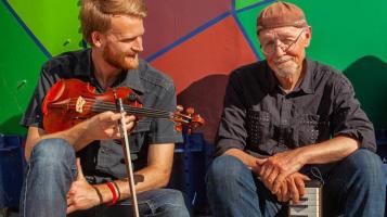 Pete and Oliver holding a violin and keyboard in front of multi-color geometric wall