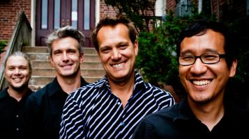 4 band members in button up shirts standing in front of brick building